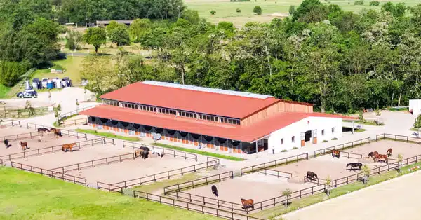 Luftaufnahme eines großen Reitstalls mit rotem Dach, umgeben von mehreren eingezäunten Paddocks, in denen Pferde grasen und sich bewegen. Die Anlage ist eingebettet in eine grüne Landschaft mit Bäumen im Hintergrund.